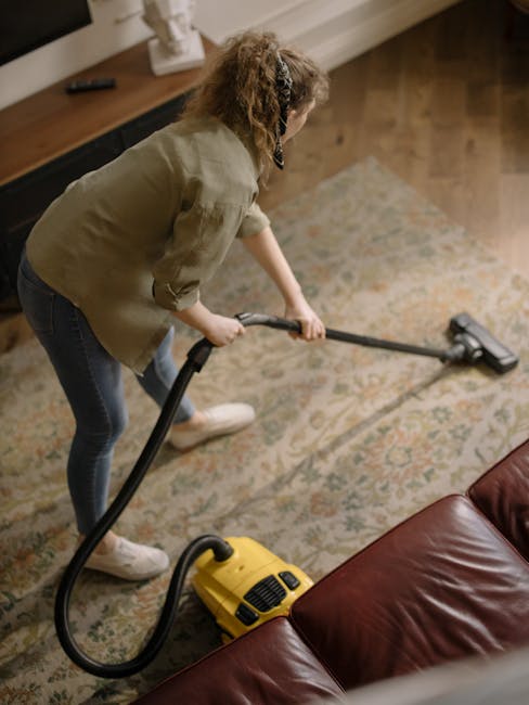 A woman with curly hair, dressed in a green jacket, blue jeans, and white sneakers, is engaged in surface cleaning using a yellow vacuum cleaner in a living room. She is holding the vacuum's hose and 