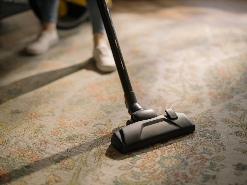 A woman with curly hair, dressed in a green jacket, blue jeans, and white sneakers, is engaged in surface cleaning using a yellow vacuum cleaner in a living room. She is holding the vacuum's hose and 